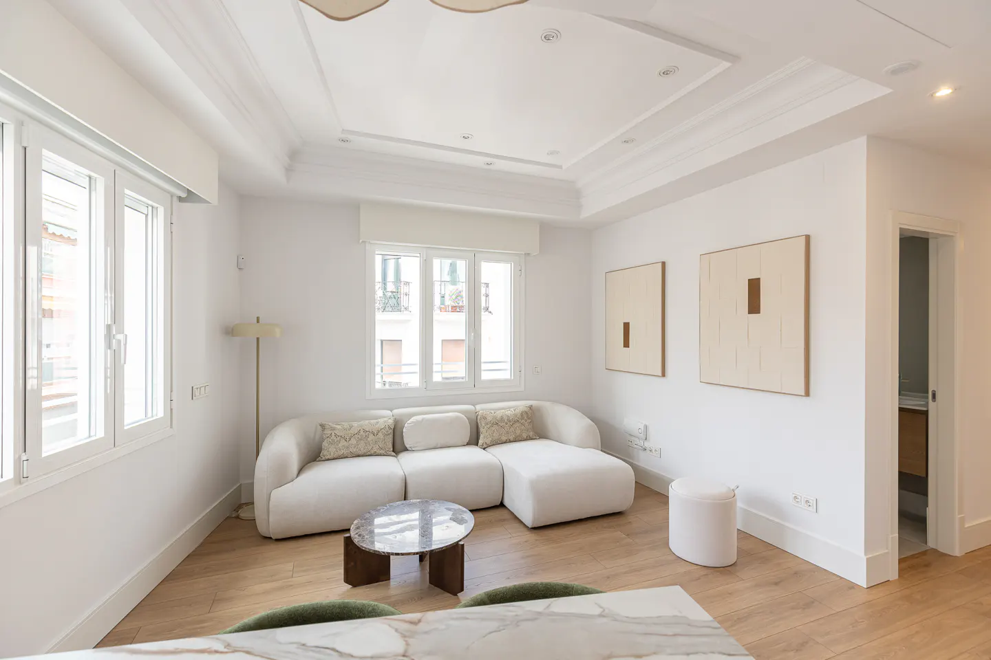 Bright living room with white walls, wood floors, and large windows. A white sectional sofa faces a marble-topped coffee table. Two framed abstract art pieces hang on the wall.