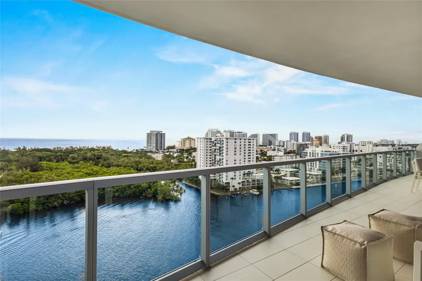 Balcony view of a city skyline, blue water, and green trees under a blue sky. Two beige cushions sit on the tiled balcony floor.