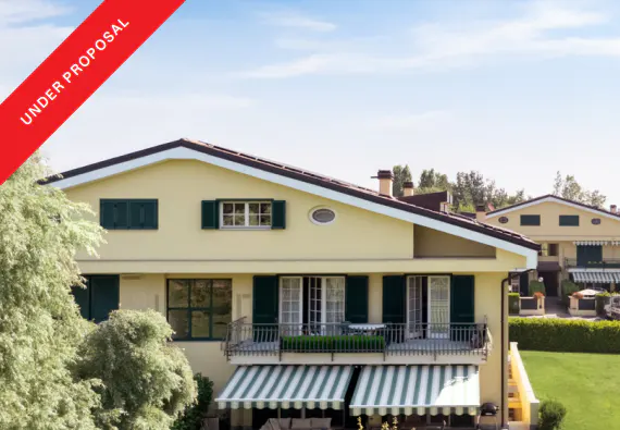 Two-story yellow house with dark green shutters and a balcony with a green and white striped awning. "Under Proposal" banner in the upper left corner.