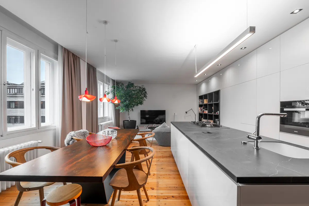A modern, open-concept kitchen and dining area with wood floors, a long wood table, and white cabinets. Red pendant lights hang above the table.