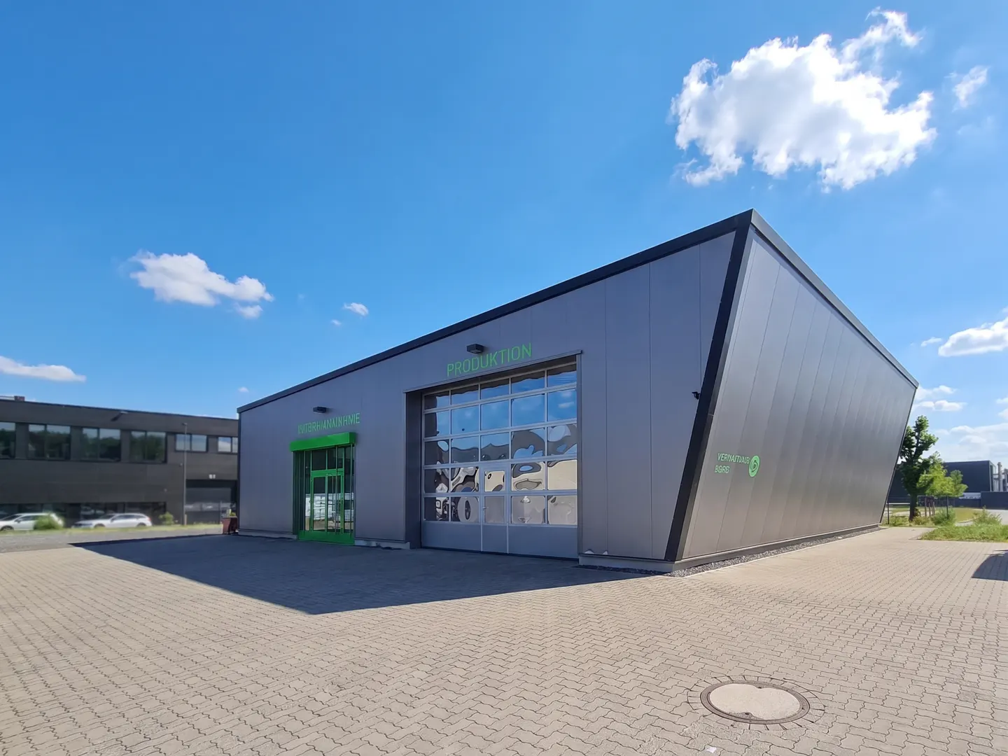 Exterior view of a gray industrial building with "PRODUKTION" and "WARENANNAHME" in green lettering, under a blue sky.