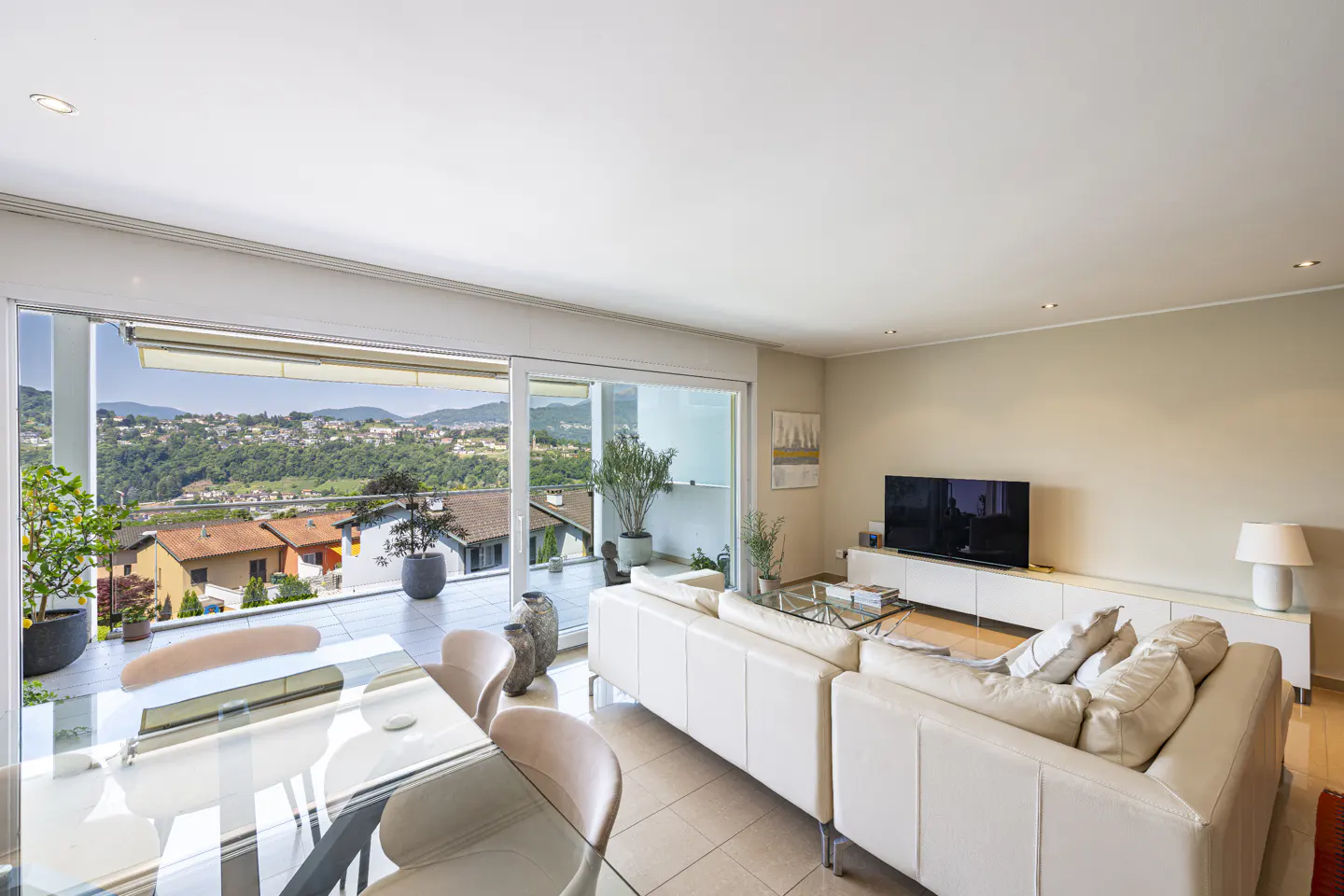 Modern living room with white sectional sofa, glass dining table, and large sliding doors opening to a scenic balcony.