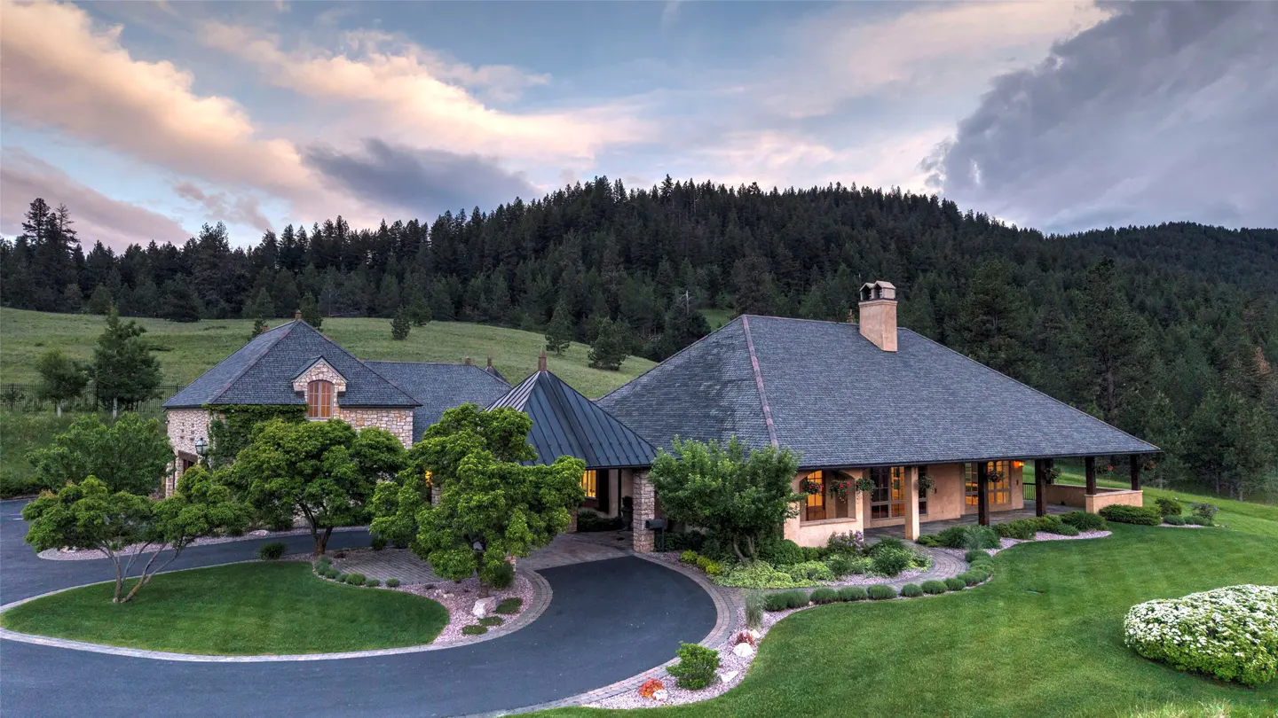 A stone house with a dark roof and circular driveway sits on a lush green lawn against a backdrop of pine-covered hills.