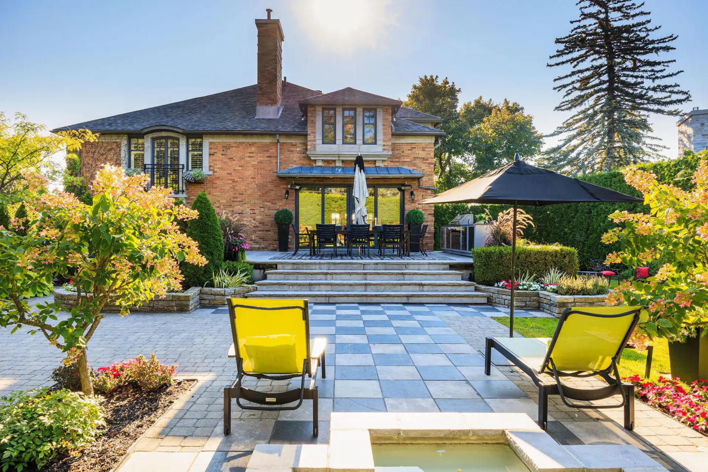 Brick house with a stone patio, lounge chairs, and a dining table. The patio has a small pool and is surrounded by greenery.