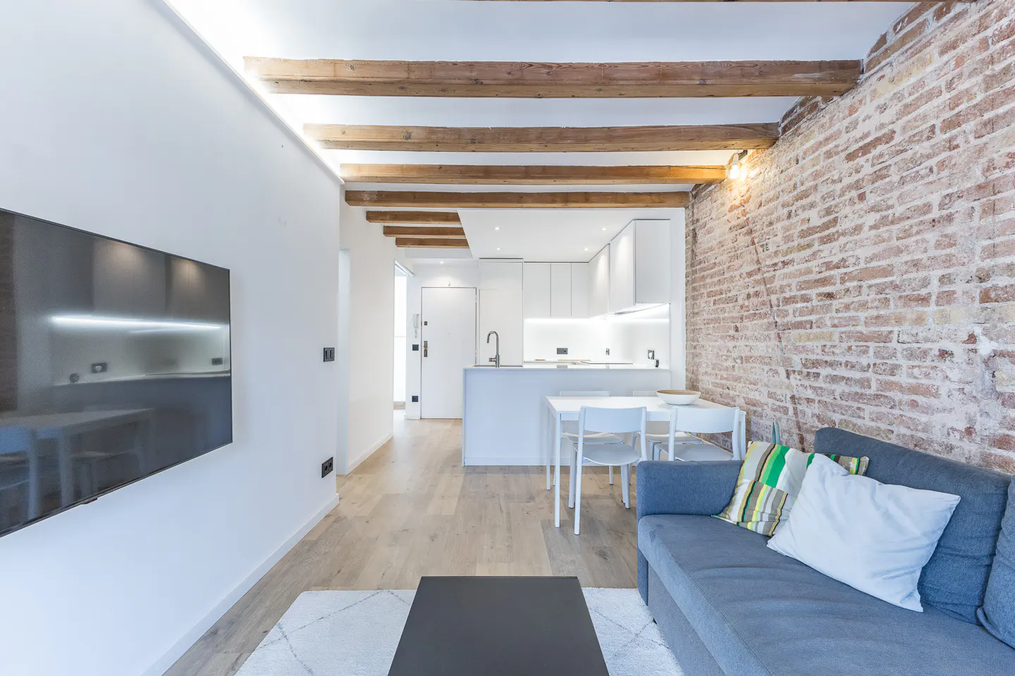 Living room with exposed brick wall, wooden ceiling beams, and a gray sofa. A modern white kitchen is visible in the background.