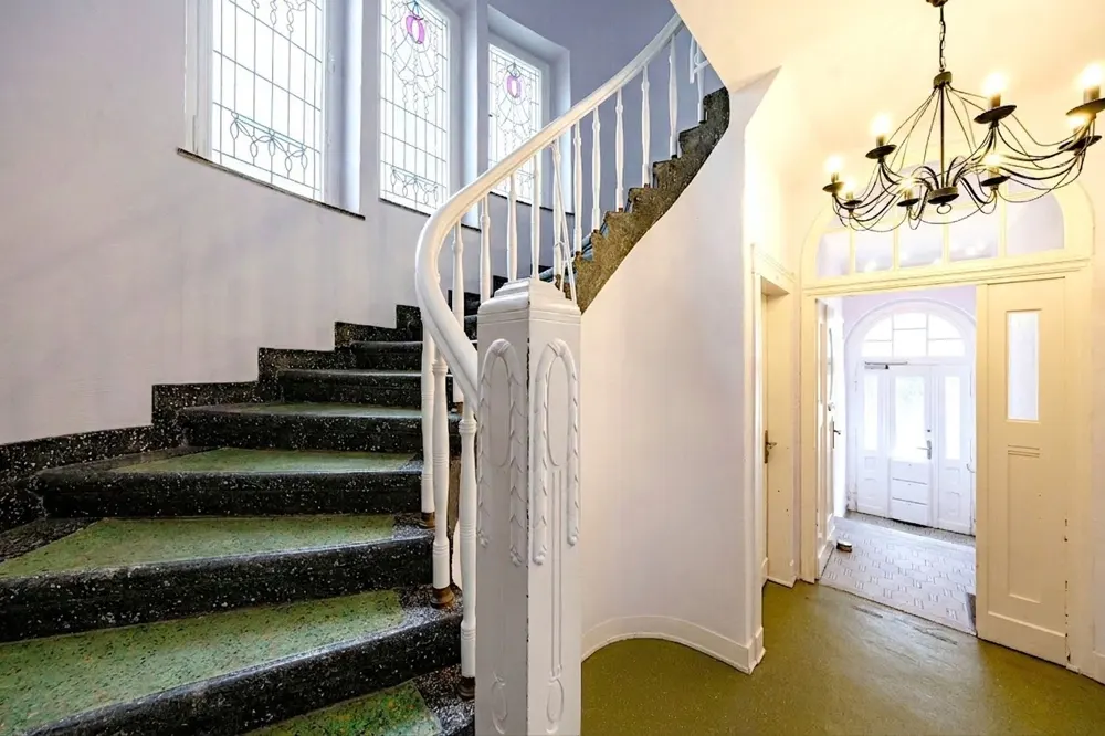 Interior view of a home's foyer with a winding staircase, white banister, and a chandelier. A doorway leads to a bright room.