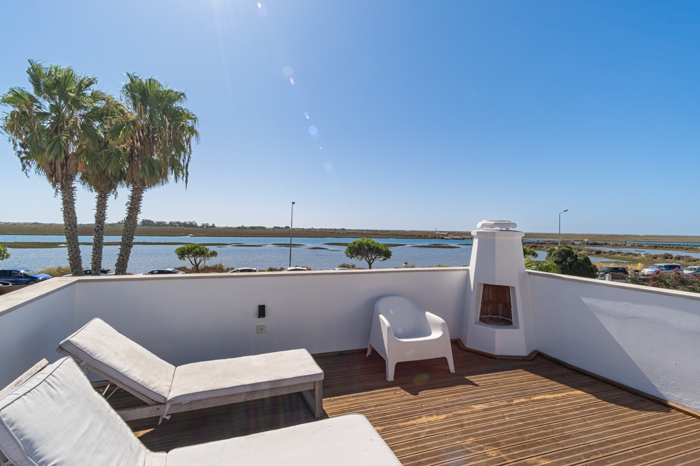 Sunny rooftop terrace with wooden decking, two white lounge chairs, a white chair, and a fireplace overlooking a lagoon. [AI-generated text]