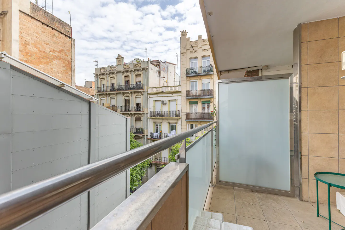Balcony view with metal railing, frosted glass panels, and tiled floor. Buildings are visible in the background under a cloudy sky.