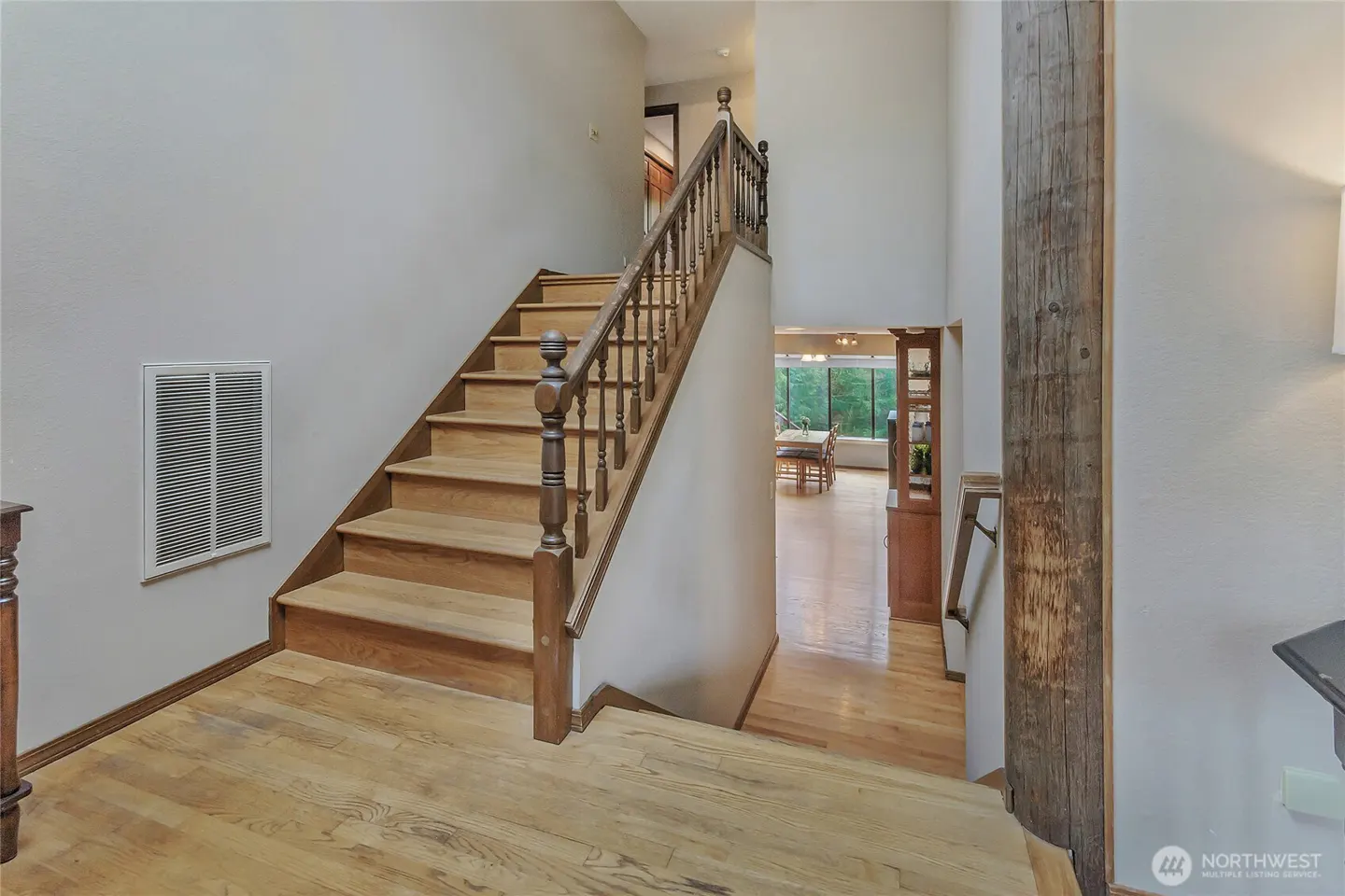 Interior view of a home featuring wooden stairs with a brown banister, light gray walls, and a glimpse of a dining room.