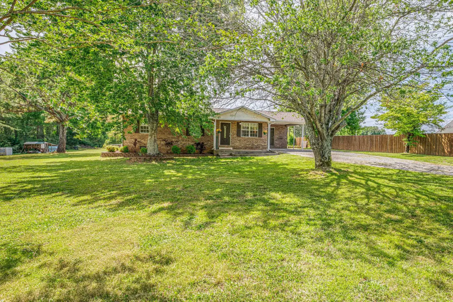 A single-story brick house with a carport, viewed from the front lawn with trees casting shadows.