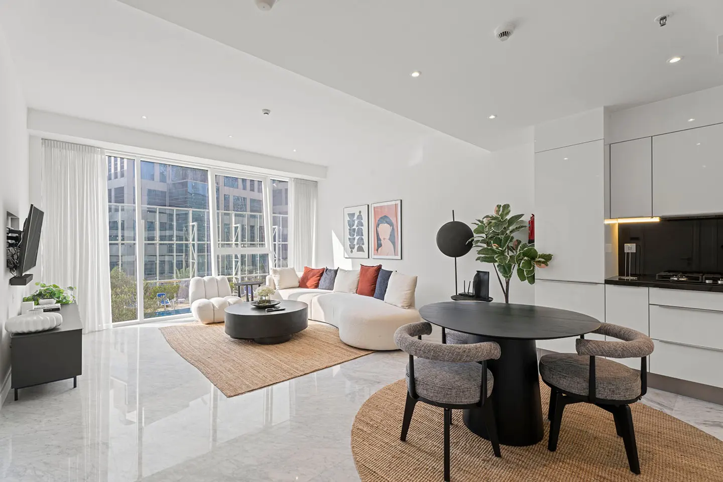 Bright, modern apartment with white walls, marble floors, and large windows. A curved white sofa faces a black coffee table. A black dining table with two chairs sits nearby.
