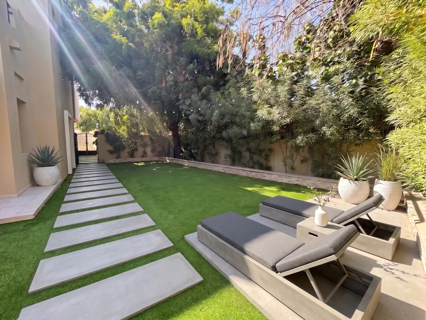 Backyard with a stone path, green lawn, two grey lounge chairs, and potted plants under lush trees on a sunny day.