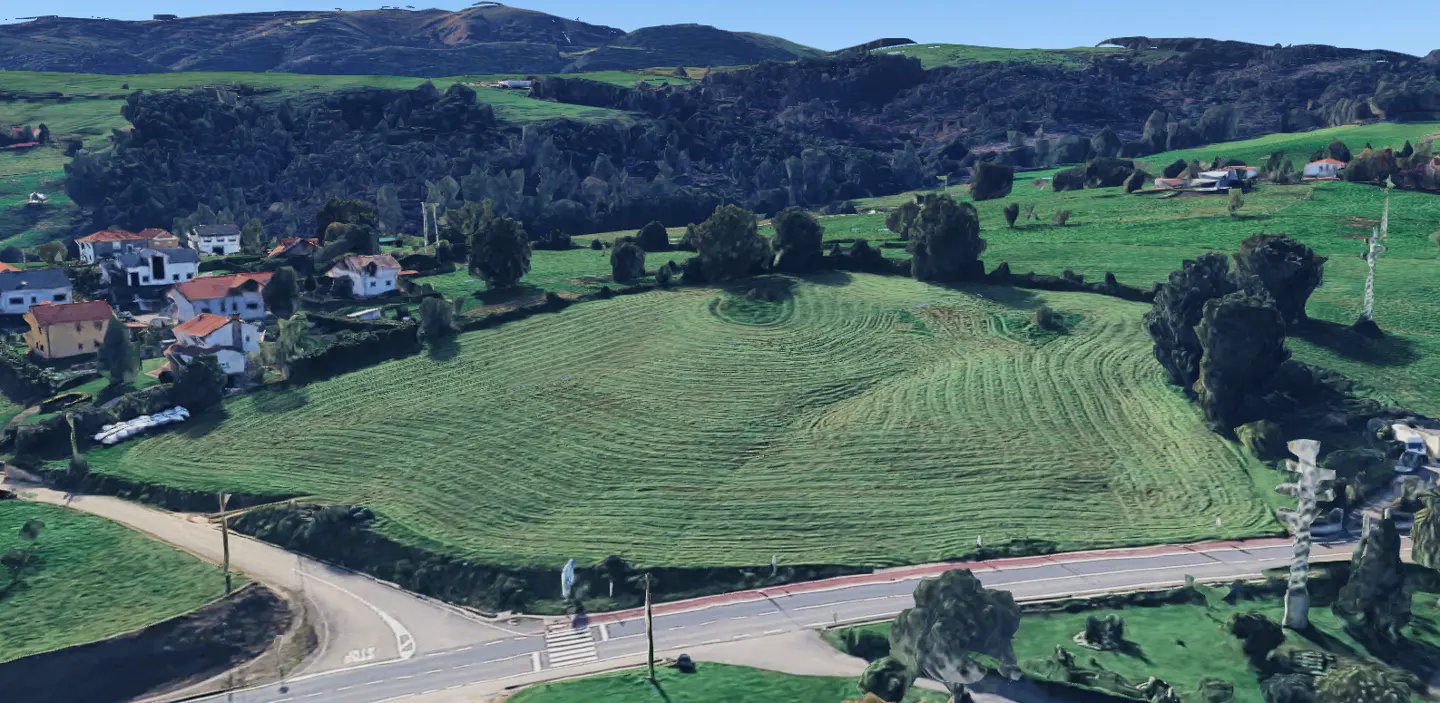 Aerial view of a large, green, grassy lot bordered by a road, trees, and houses in a rural area.