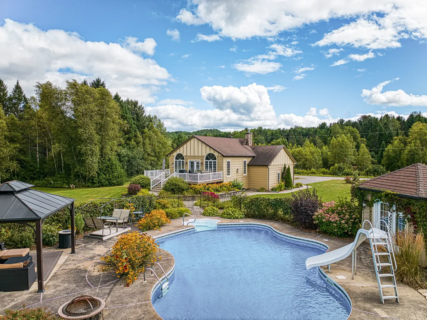 Aerial view of a kidney-shaped pool with a slide, a gazebo, and a yellow house with a white deck surrounded by green trees.