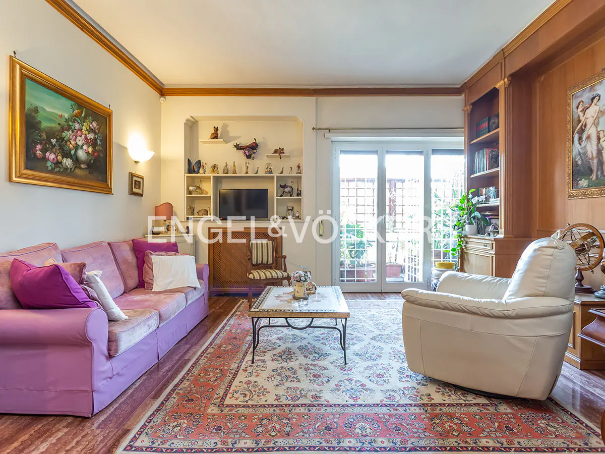 Living room with a purple sofa, white armchair, patterned rug, and a coffee table. Shelves with decor and a TV are in the background.