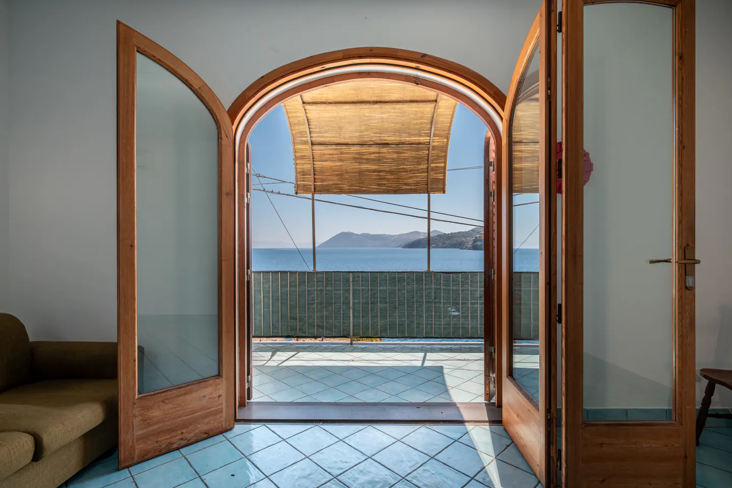 Open arched wood doors lead to a balcony with ocean views. Blue tiled floor, woven shade, and green railing visible.