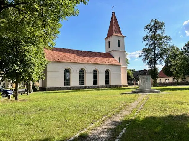 A white church with a red roof and steeple stands on a green lawn under a blue sky. A stone monument is in front of the church.