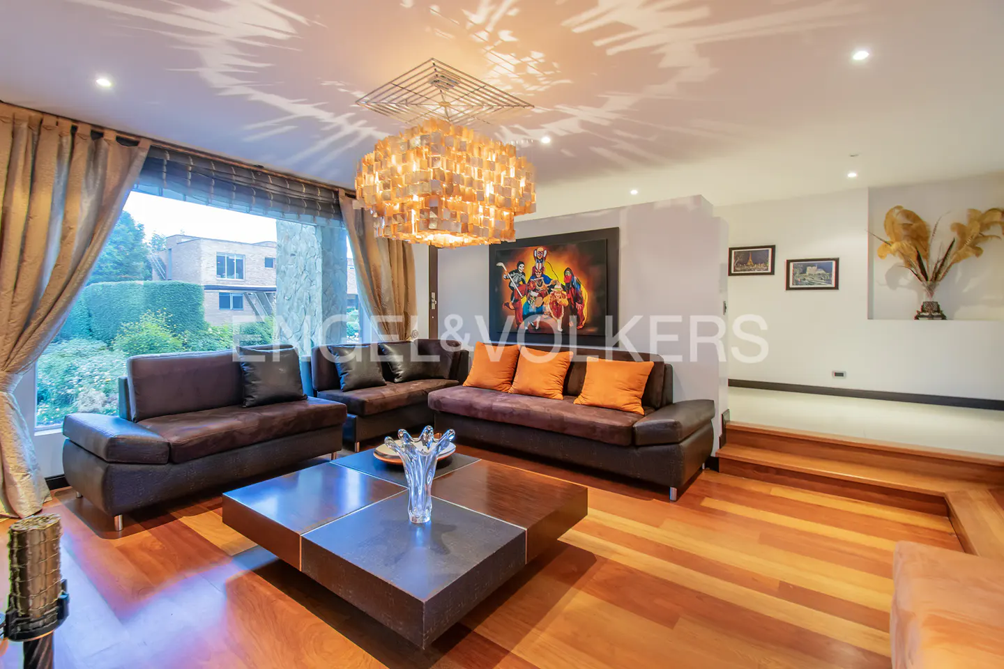 Living room with brown sofas, orange pillows, wood floors, and a large window with curtains. A modern chandelier hangs from the ceiling.