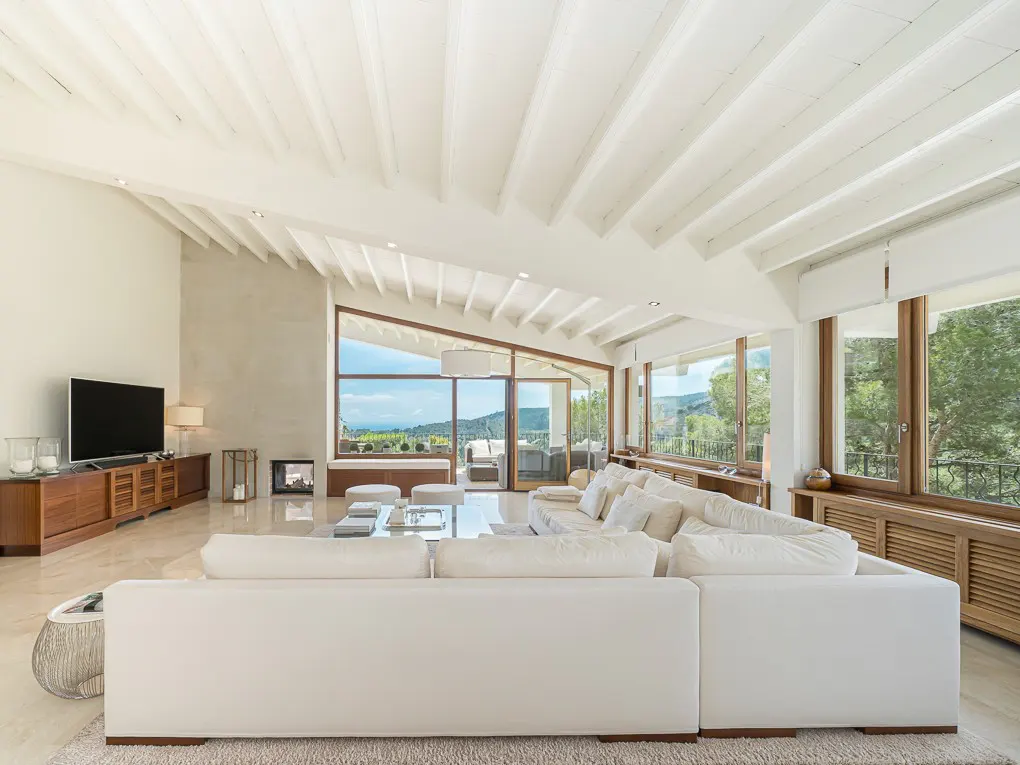 Bright living room with white sofa, wood cabinets, and large windows overlooking a green landscape.