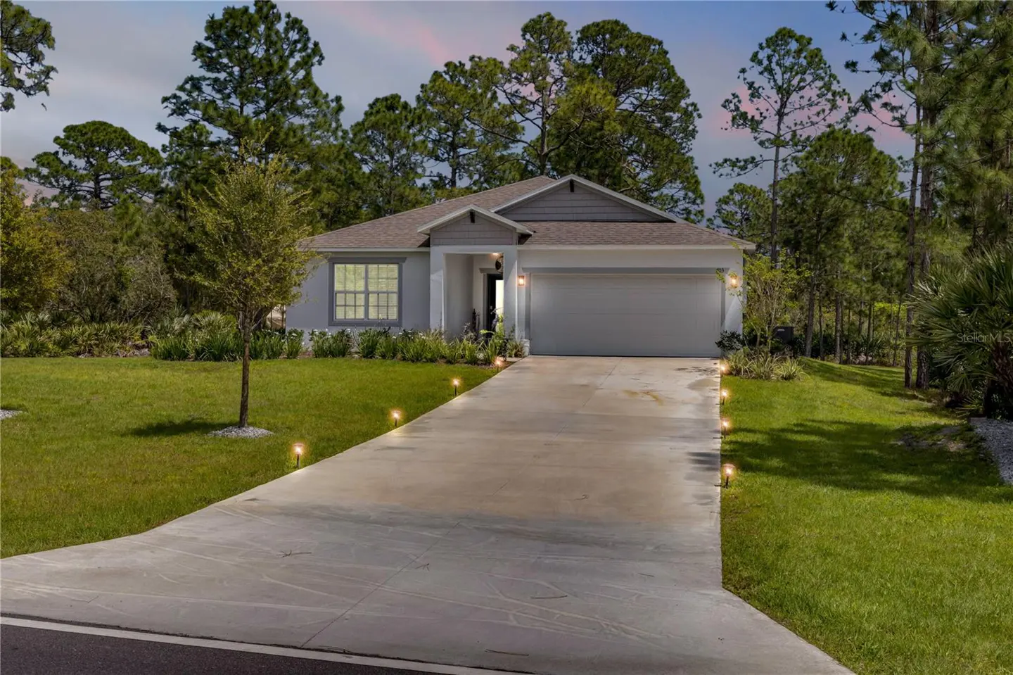 Single-story gray home with a two-car garage, concrete driveway, and landscape lighting at dusk, surrounded by pine trees.