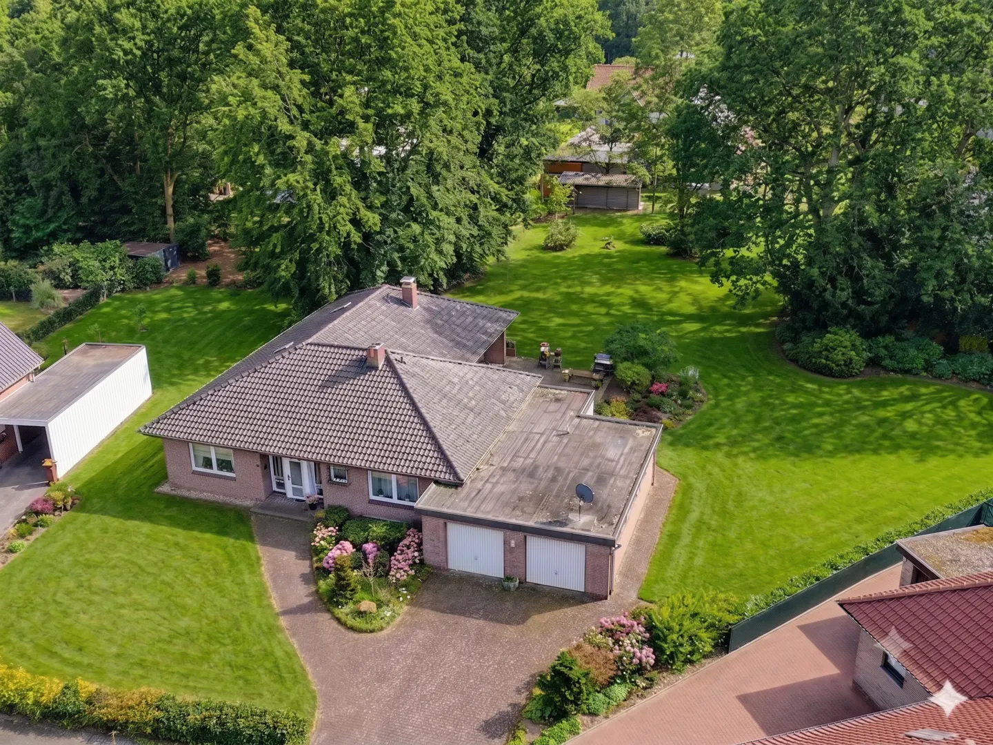 Aerial view of a brick house with a tiled roof, double garage, and a large, lush green backyard surrounded by trees.