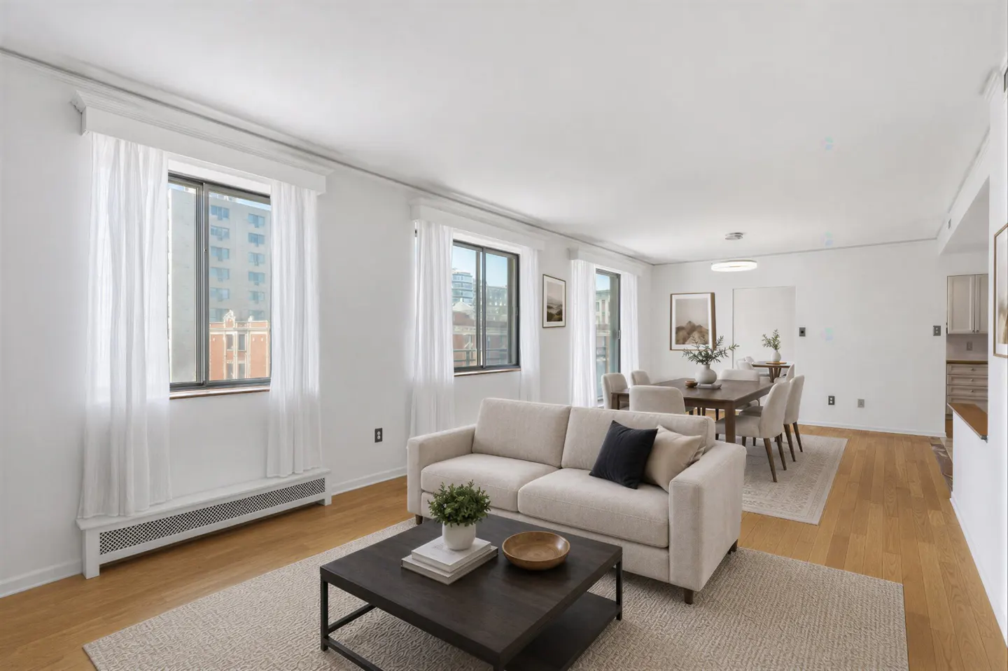 Bright living room with hardwood floors, white walls, and large windows with sheer white curtains. A beige sofa faces a dark wood coffee table. Dining table in background.