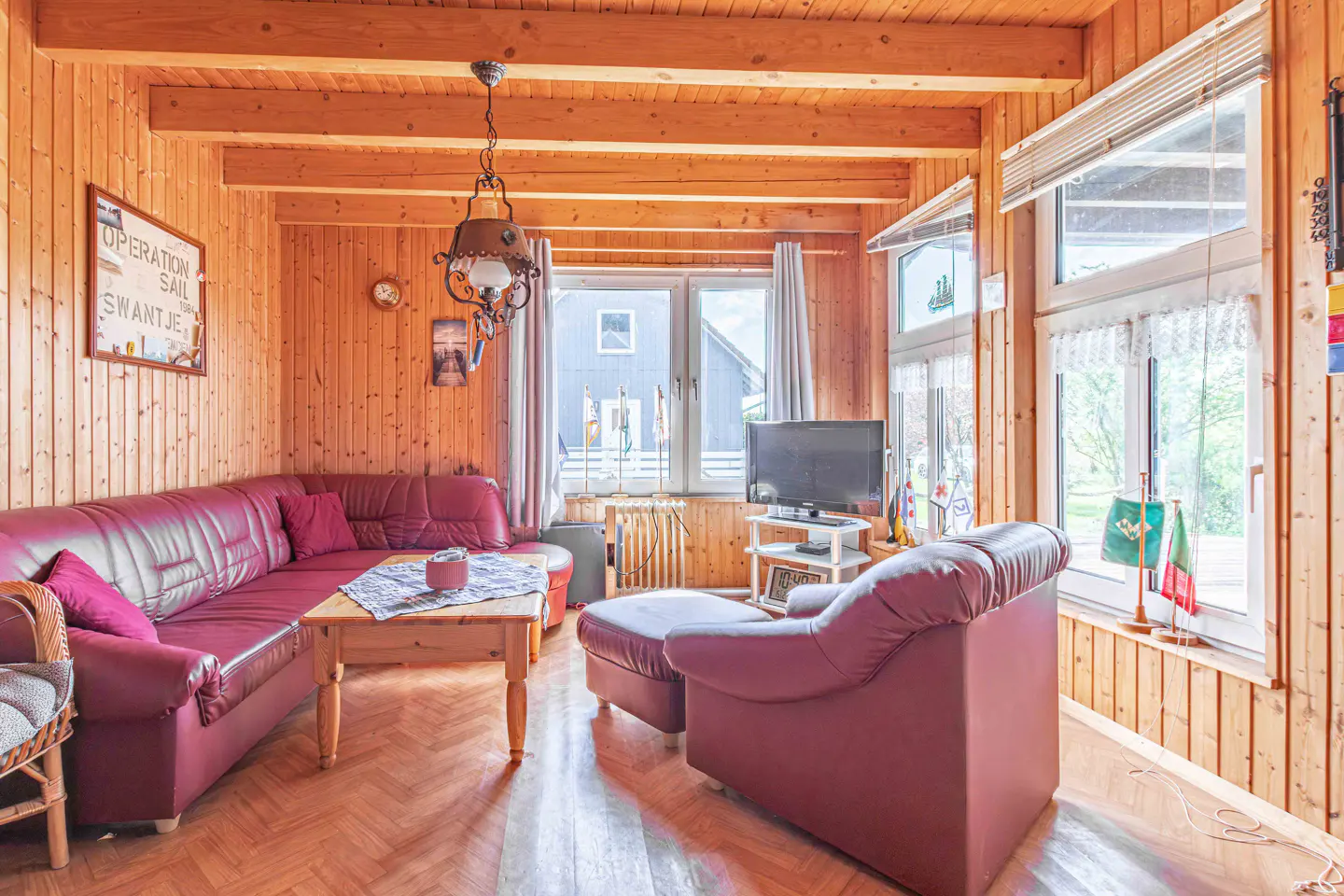 Cozy living room with wood-paneled walls, red leather sofa, armchair, and a wooden coffee table. Large windows offer natural light.
