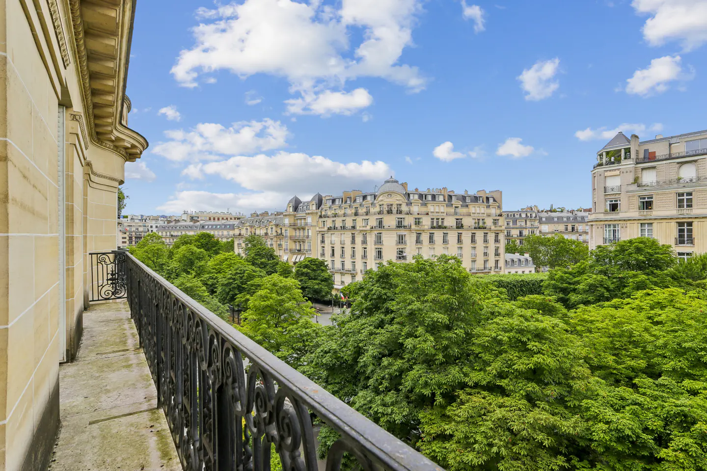 View from a stone balcony with a black iron railing overlooking lush green trees and classic Parisian apartment buildings.