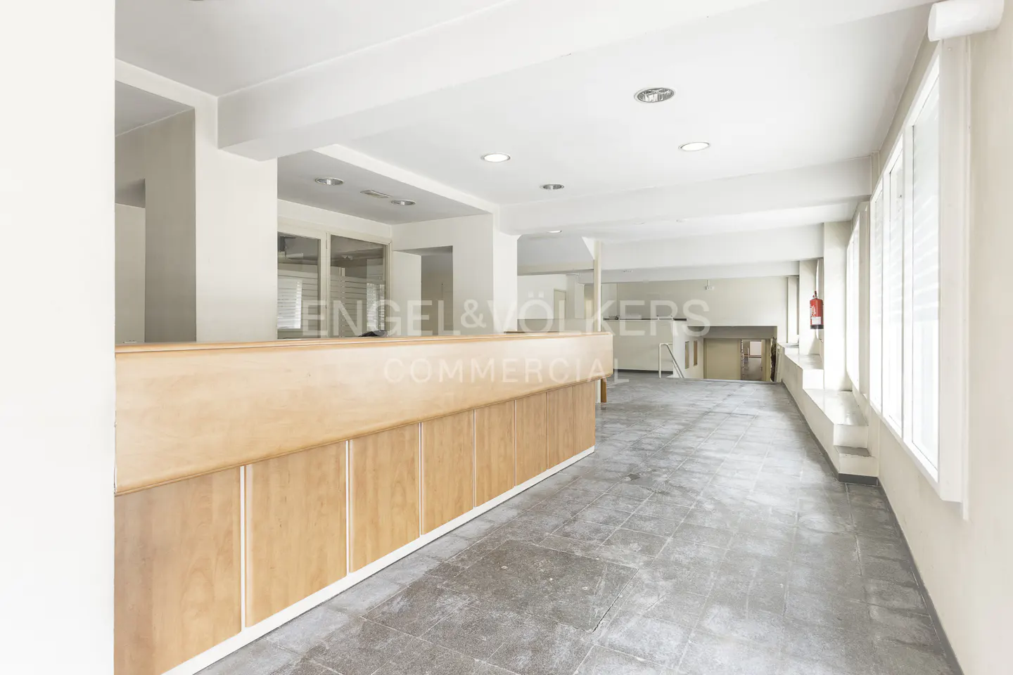 Empty commercial space with a light wood reception desk, gray tile floor, white walls, and large windows.