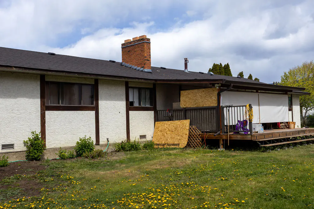 Exterior view of a one-story house with a brick chimney, dark roof, and a wooden deck. The lawn is green with yellow dandelions.