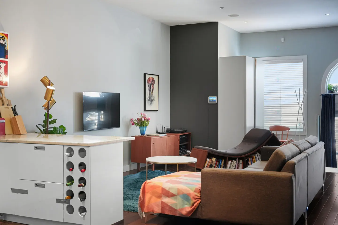 Open-concept living room with a brown sofa, chaise lounge, and white kitchen island with wine storage. A TV hangs on the gray wall.