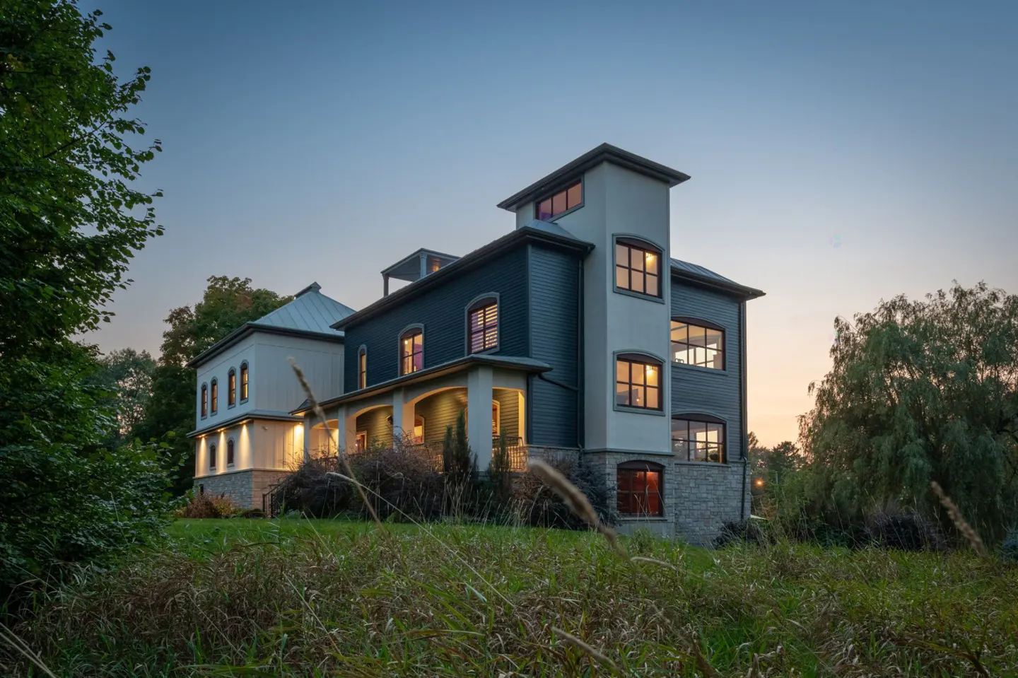 Exterior view of a large, two-story house with a tower, set against a twilight sky and grassy foreground.