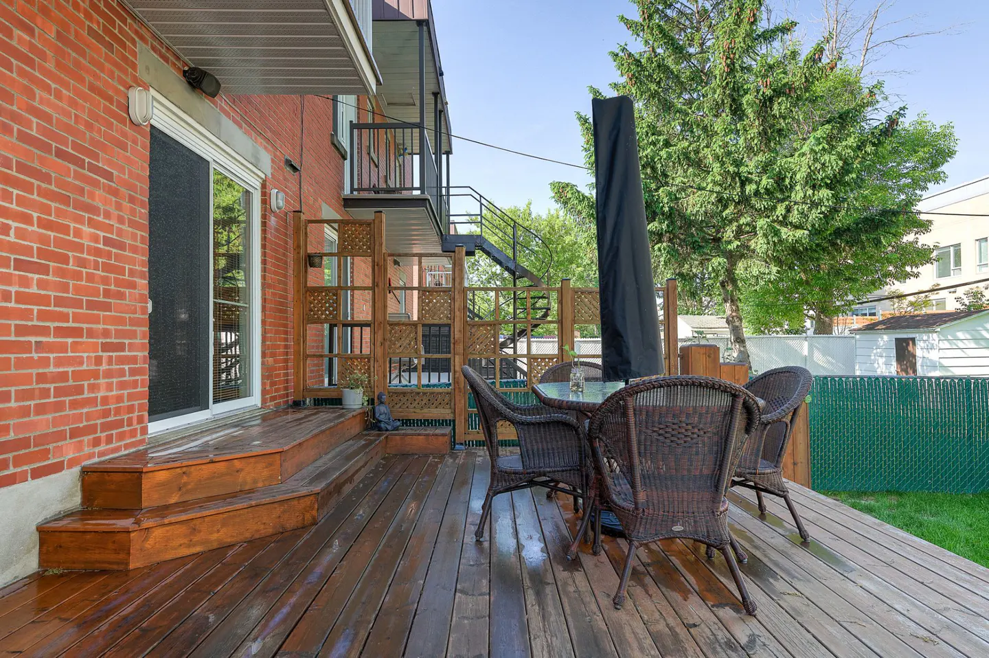 A wooden deck with a table and chairs is next to a red brick building. A black umbrella is folded up next to the table.