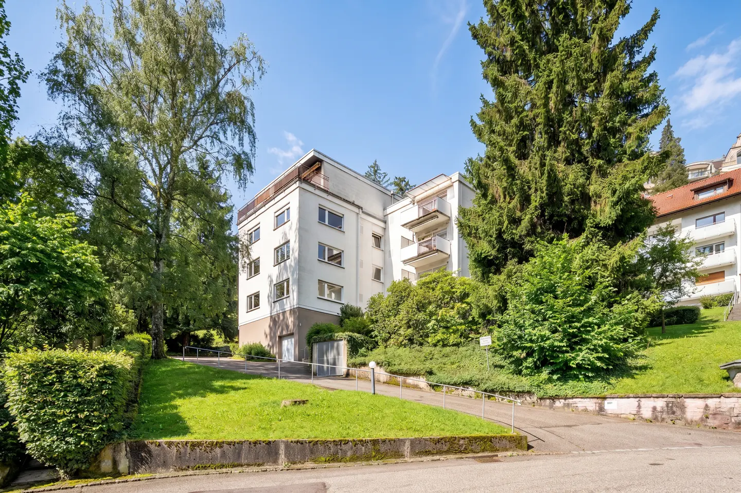 Exterior view of a white, multi-story apartment building with balconies, surrounded by lush green trees and landscaping.