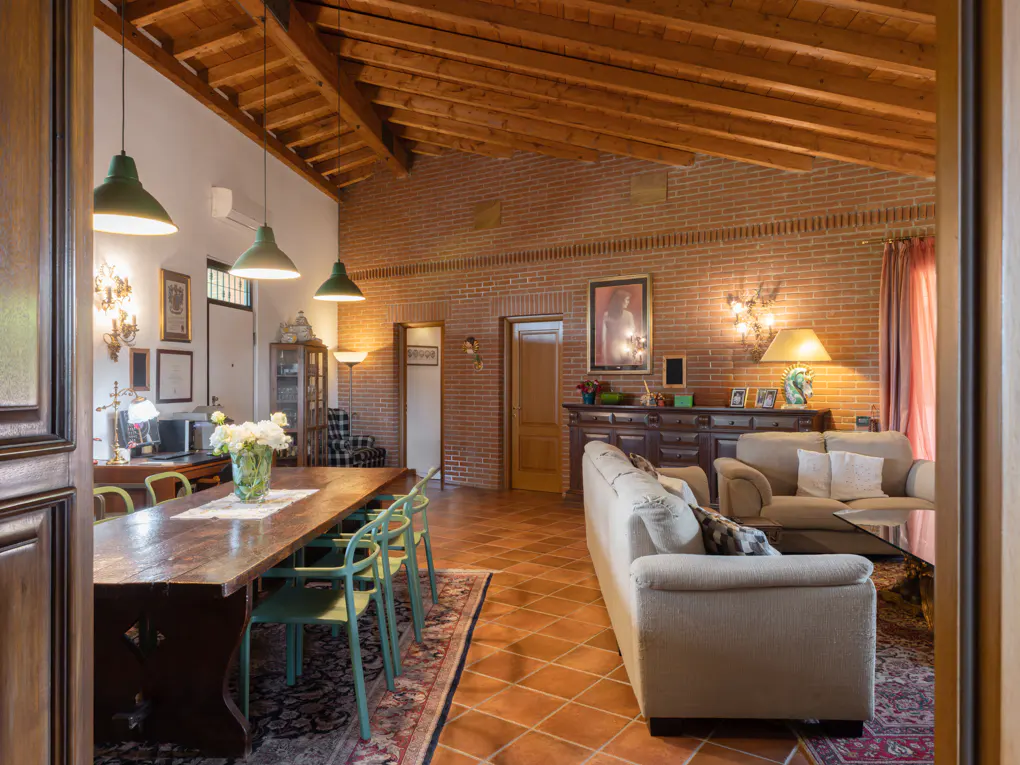 Spacious living area with exposed wood beams, brick walls, terracotta floors, a long dining table, and a cozy sofa.