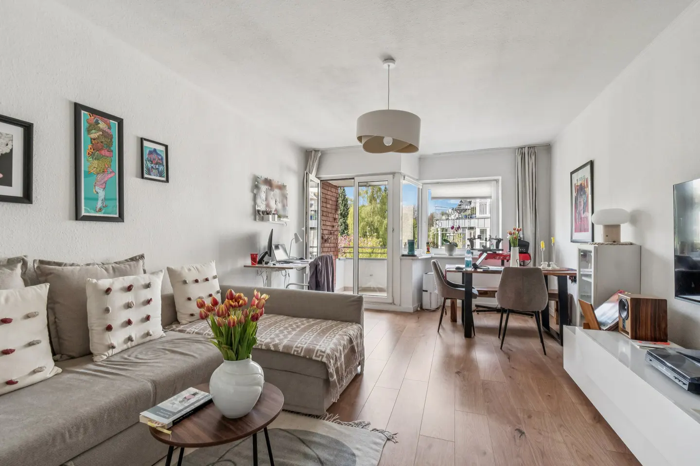 Bright living room with hardwood floors, a gray sofa with decorative pillows, and a round coffee table with tulips. A desk and dining area are visible in the background.