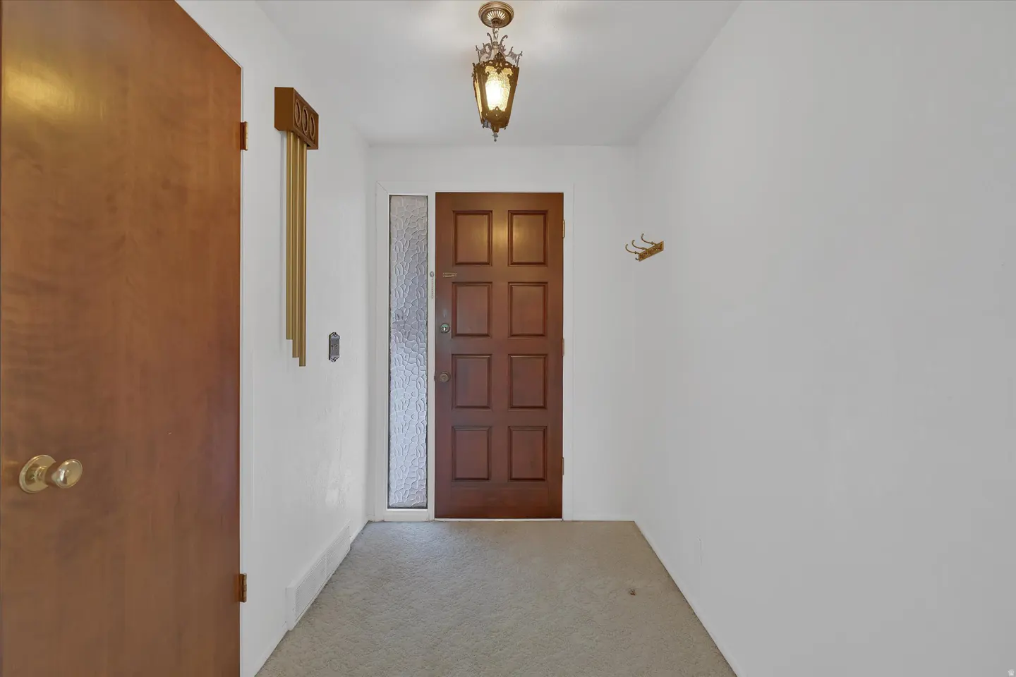Entryway with a brown front door, white walls, and gray carpet. A vintage light fixture hangs from the ceiling.