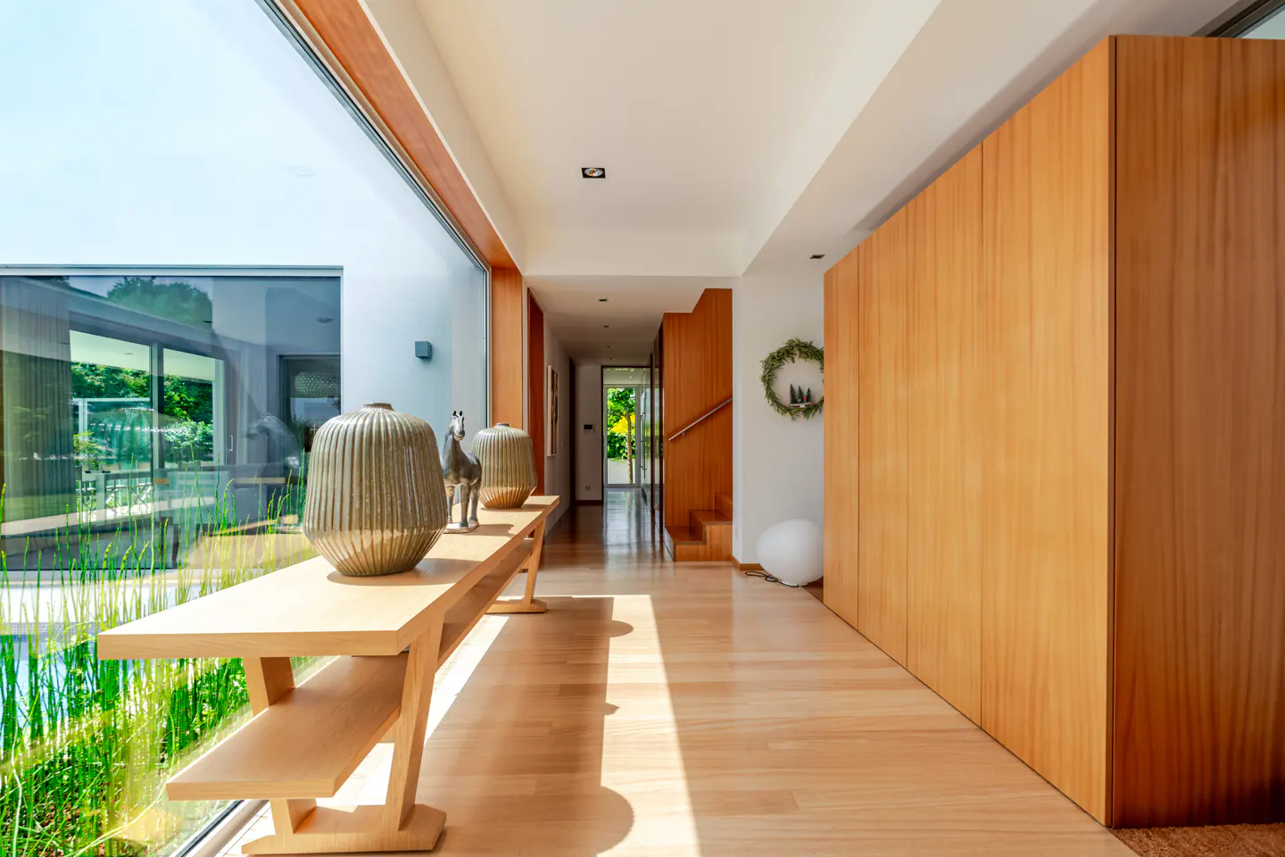 Bright hallway with wood floors, a long table with vases, and a large window overlooking greenery. A wooden staircase and closet are visible.