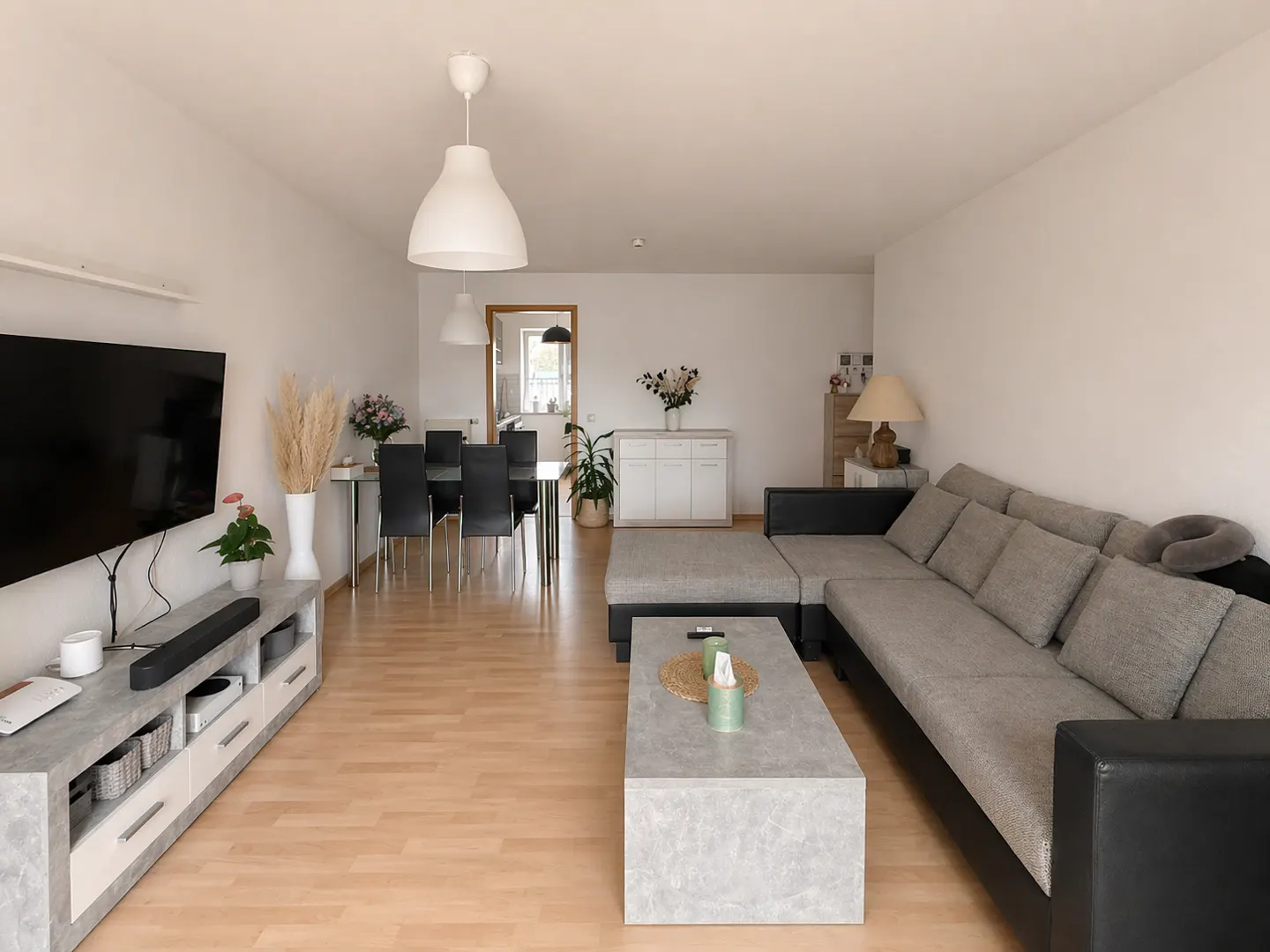Bright living room with a gray sectional sofa, marble coffee table, TV, and dining area with black chairs.
