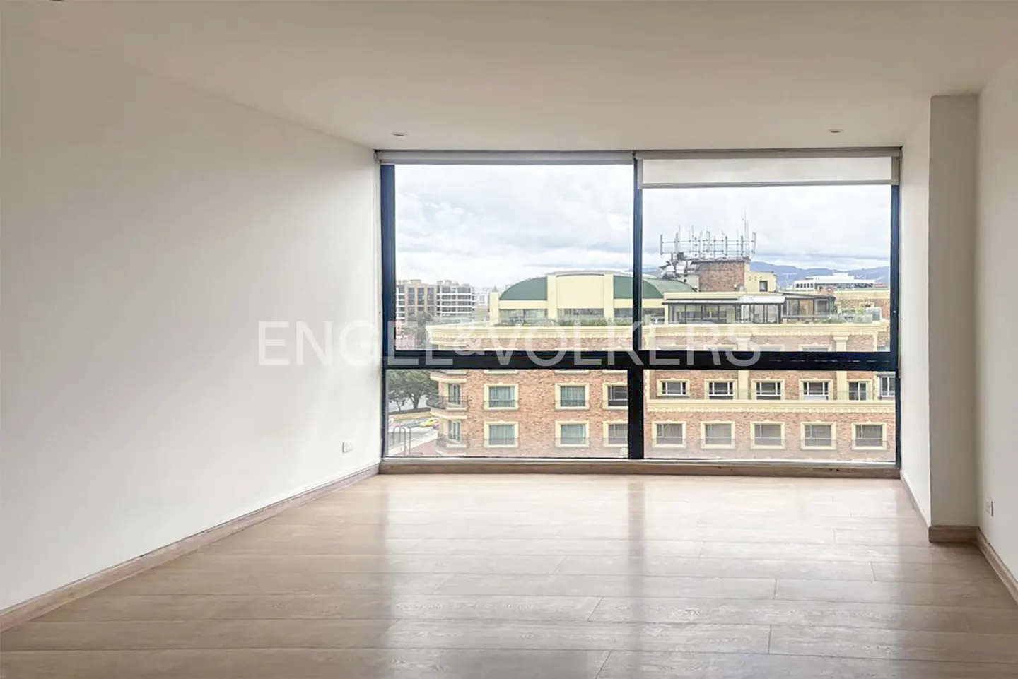 Empty room with light wood floors and white walls. Large windows offer a city view.