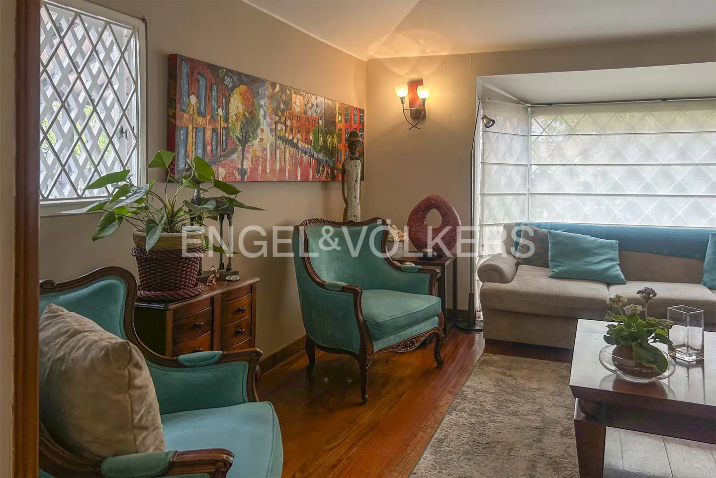 Living room with teal armchairs, a beige sofa, and a dark wood coffee table. A colorful painting hangs above a wooden dresser.