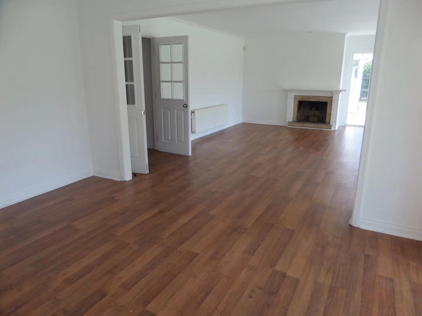 An empty living room with wood floors, white walls, a fireplace, and a door leading to another room.