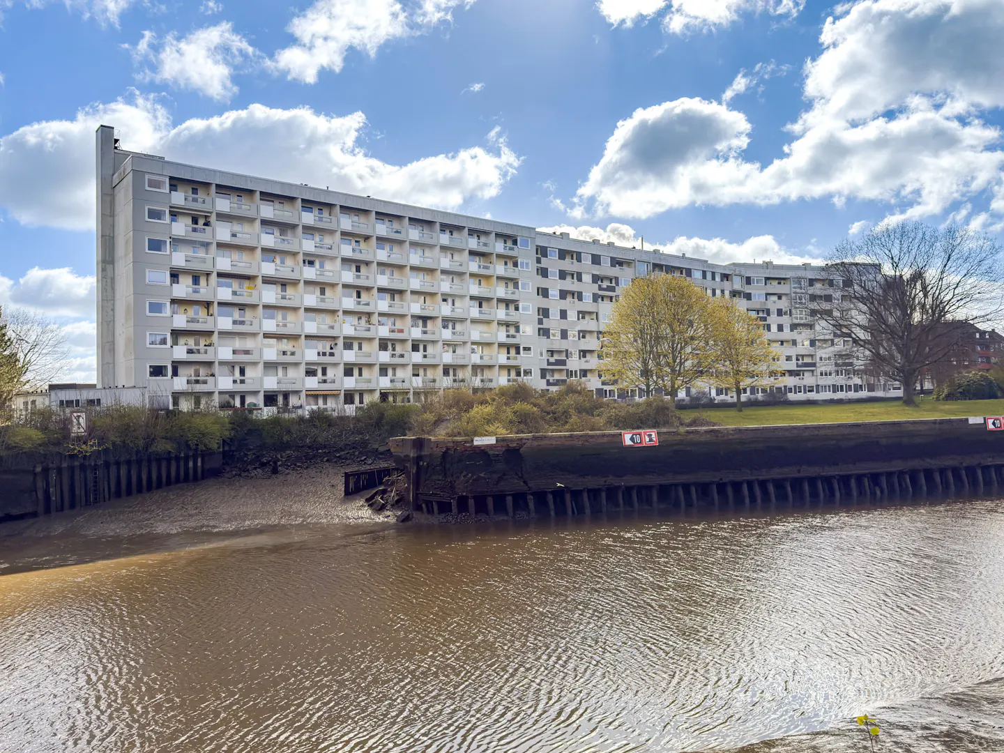 Exterior view of a large, gray apartment building with balconies, situated along a muddy riverbank under a blue sky with white clouds.