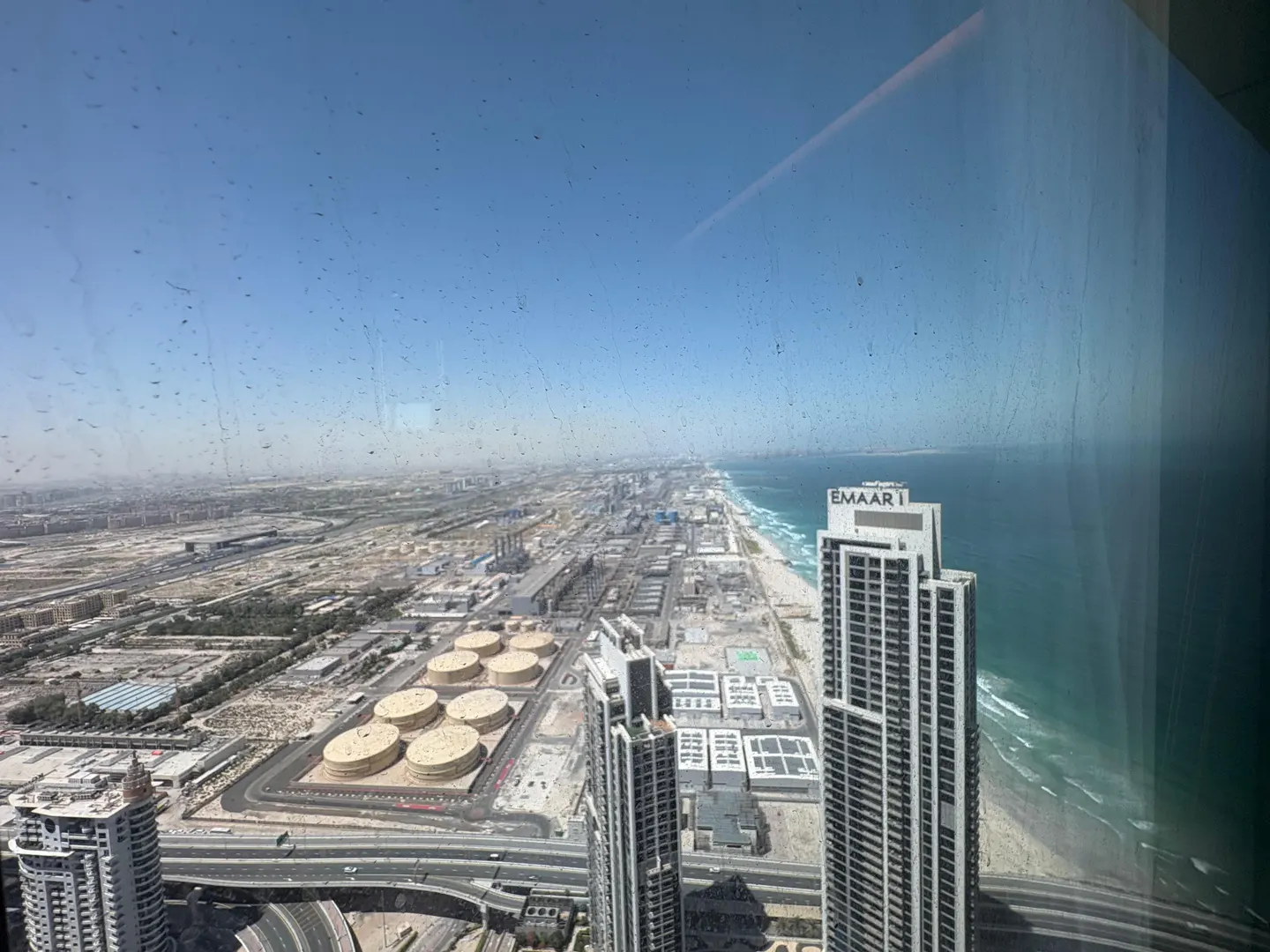 High-angle view through a window showing Emaar skyscrapers, industrial tanks, and the coastline under a clear blue sky.