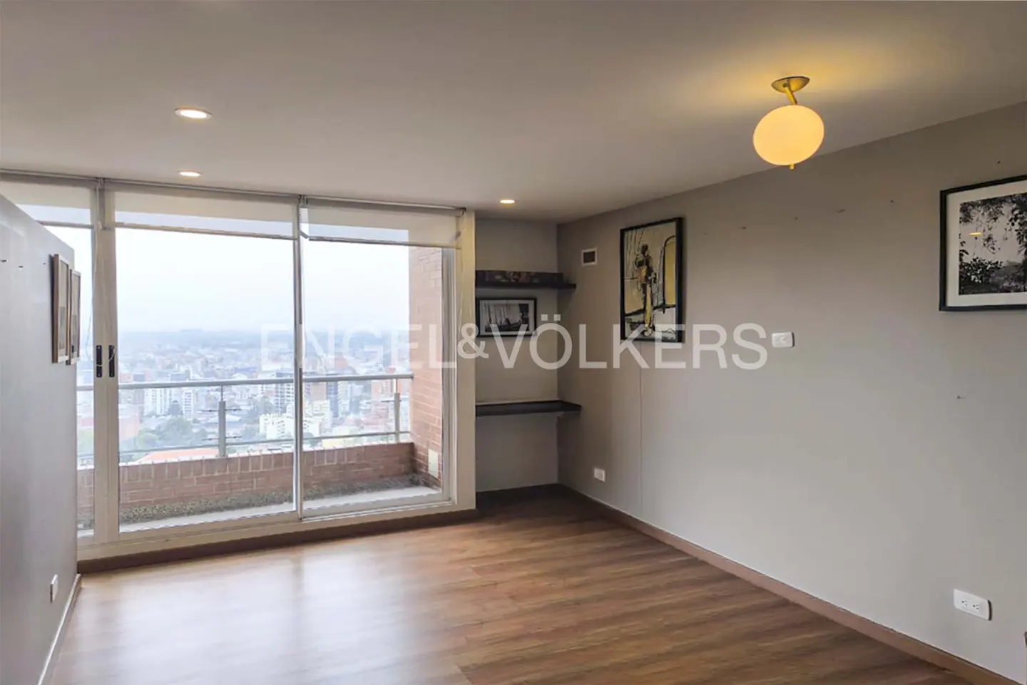 A bright, empty room with wood floors, gray walls, and a balcony overlooking a city. Artwork hangs on the walls.