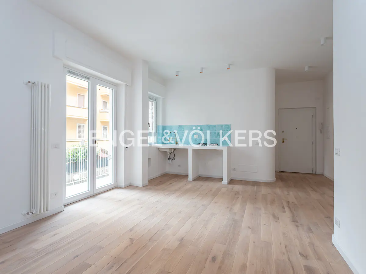 Bright, empty apartment with light wood floors, white walls, and a small kitchen area with blue tile backsplash. Balcony doors open to a view of a building.