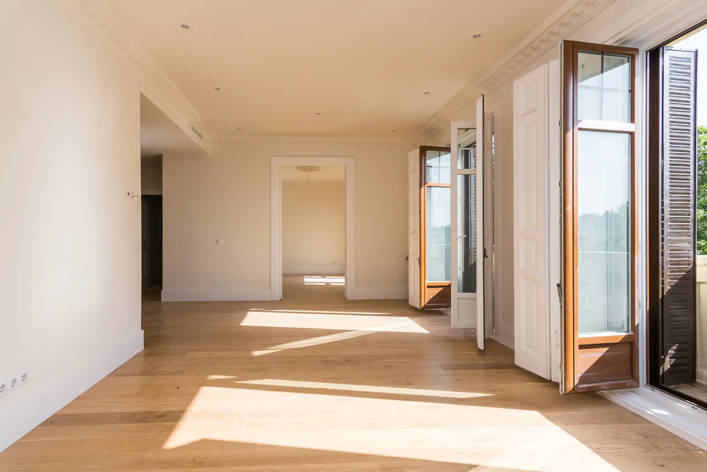 Empty room with light wood floors, cream walls, and open French doors leading to a balcony, bathed in natural sunlight.