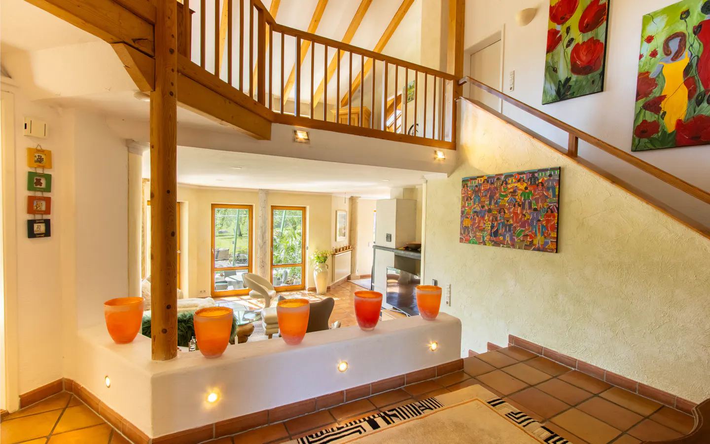 Interior view of a home with a staircase, wooden beams, and orange vases on a white ledge. Paintings adorn the walls.