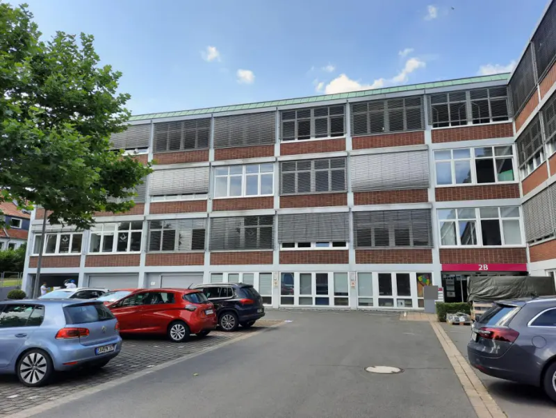 Exterior view of a three-story brick building with many windows and gray blinds. Cars are parked in front of the building. A tree is on the left.
