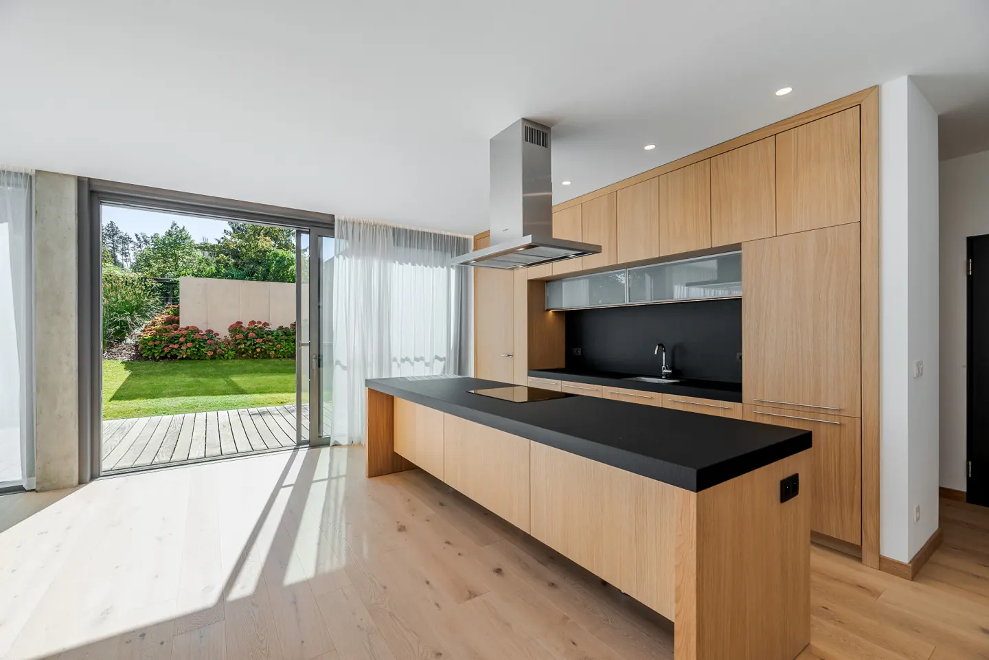 A modern kitchen with light wood cabinets, a black countertop island, and a stainless steel range hood. Sliding glass doors open to a green lawn.